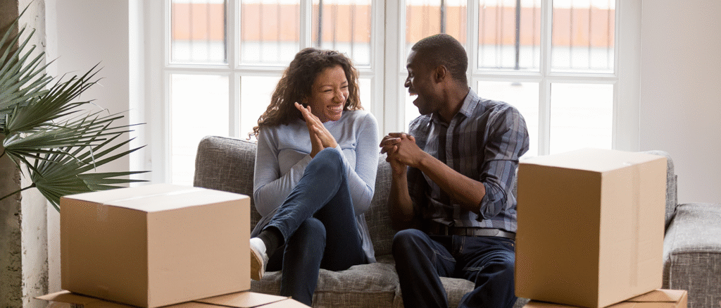 Happy young couple sitting on sofa surrounded by moving boxes in their new rental flat